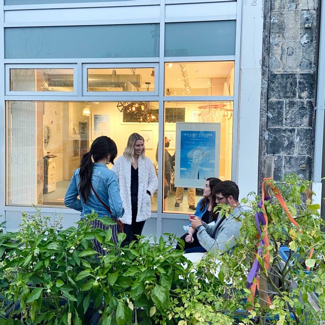 Four people talk outside a building surrounded by pots of plants.