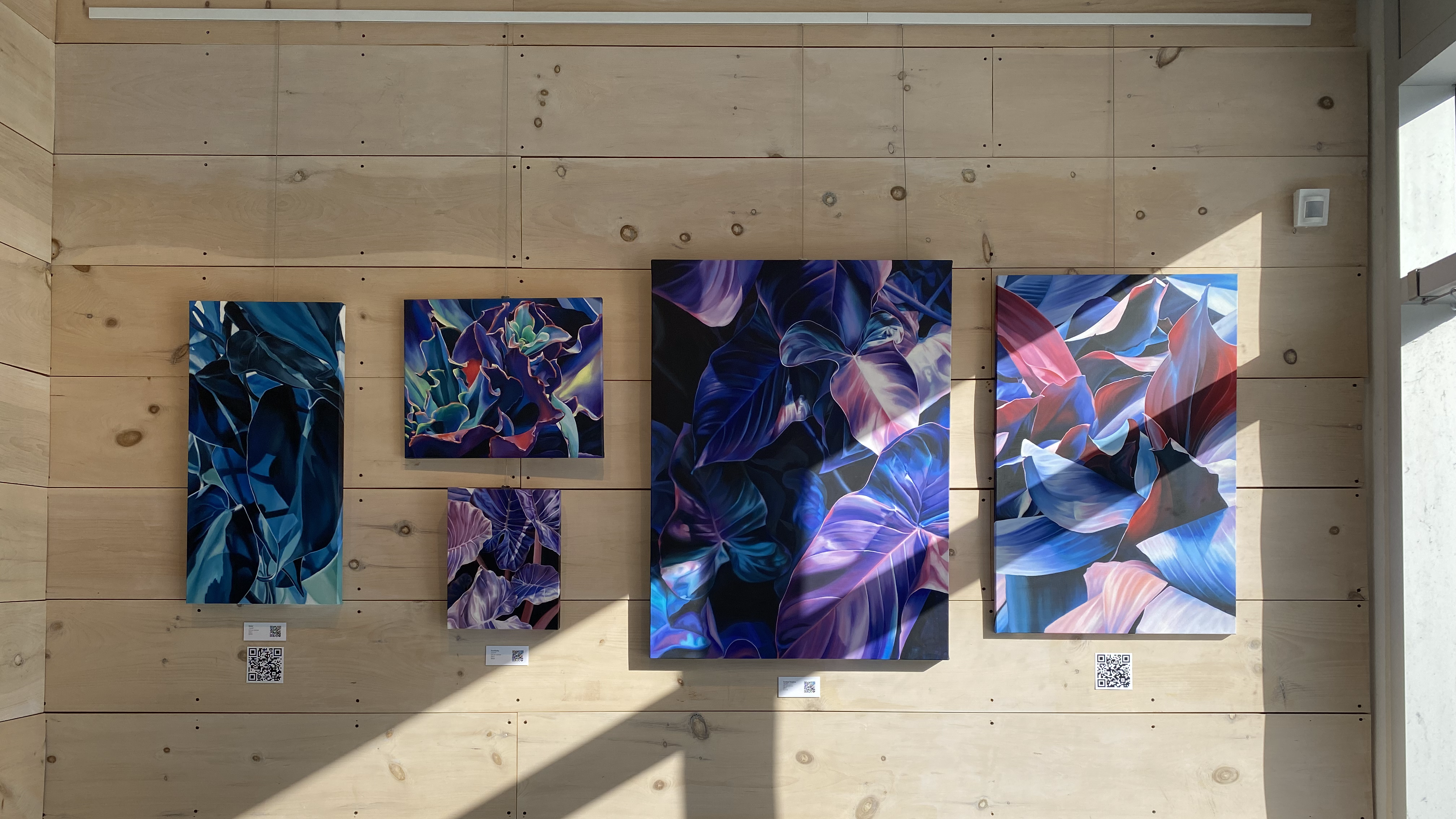 A series of five brilliant blue and purple paintings of leaves and botanical foliage are hung on a wood slat wall. The sun streams in onto the paintings through the window on the right-hand side of the photograph.