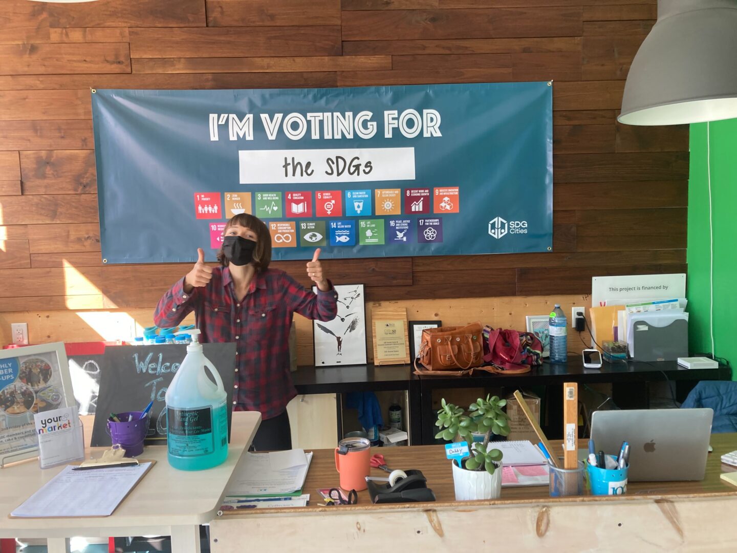 Person giving a thumbs up behind a receiption desk in front of a banner reading 'Vote for the SDGs'.