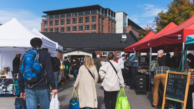 The image is focused on two people walking away from the camera holding bags, amongst other people shopping at a farmers' market with outdoor tents.