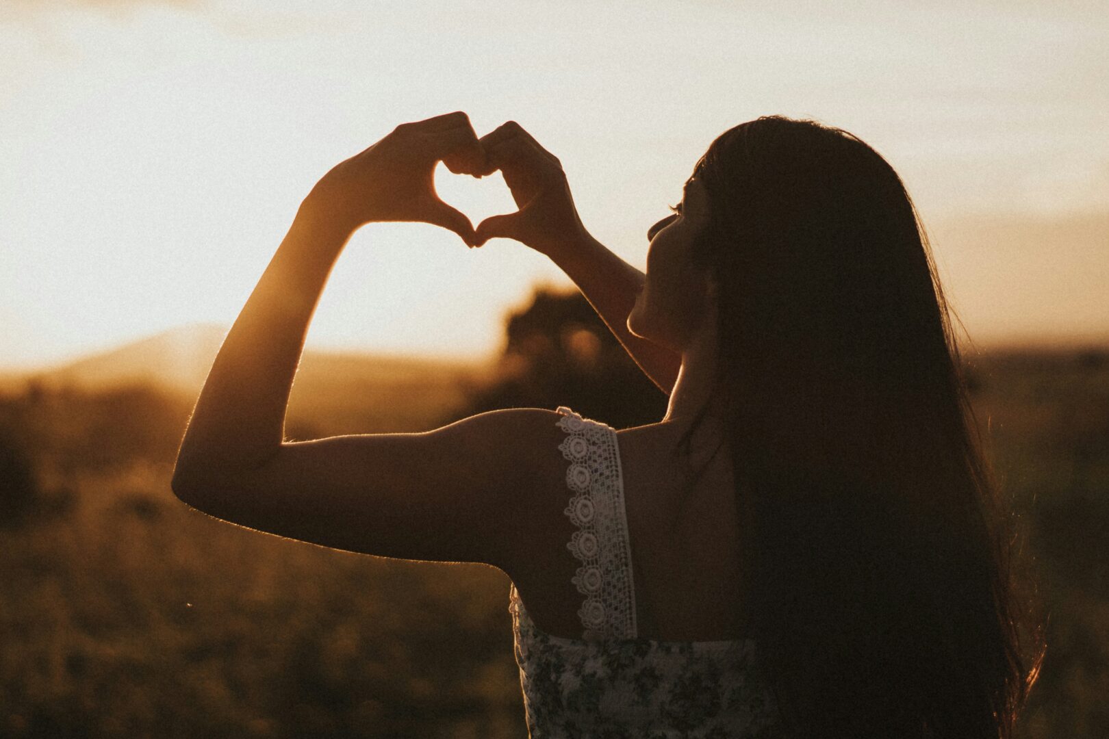 Woman in a white lace outfit making a heart shape with her hands against a sunset. 