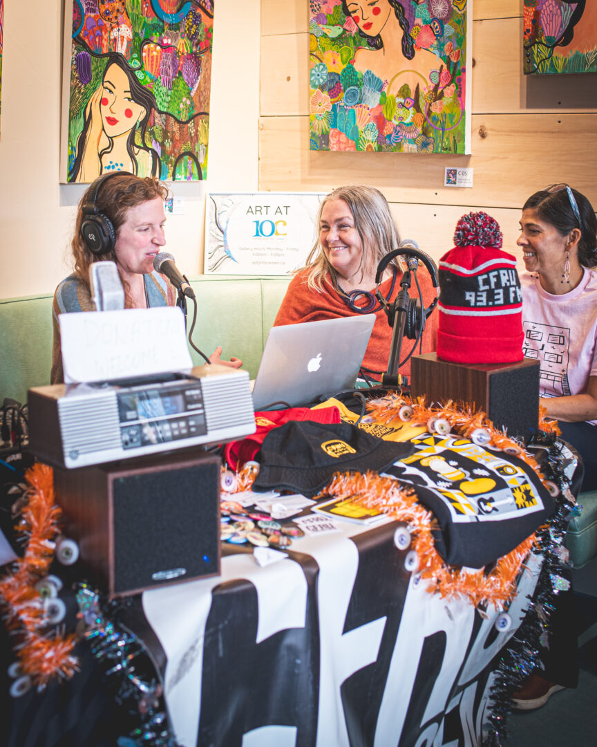 Three women smiling sitting at a table recording a radio show. Behind them colourful artwork us hung on the walls.