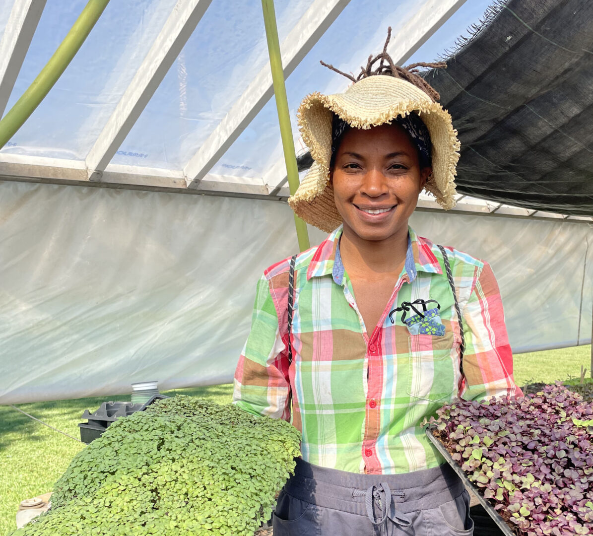 A farmer in a wide brimmed  hat smiling at the camera holding trays of micro greens