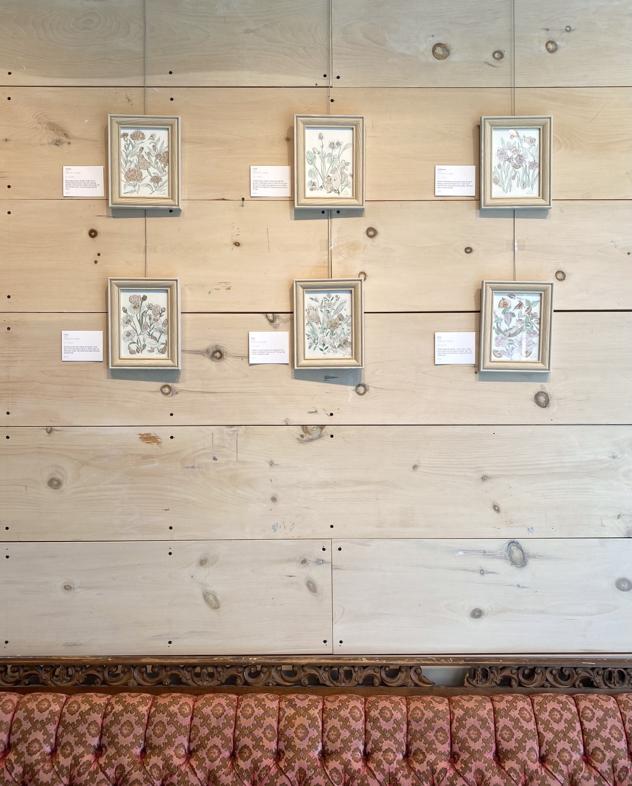6 framed watercolour artworks hanging above a red couch in the gallery